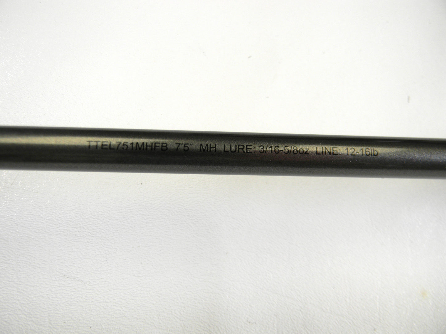 Close-up of a Daiwa Tatula Elite TTEL751MHFB 7'5" MH black fishing rod, with "LURE: 3/16-5/8oz LINE: 12-16lb" engraved. Perfect for upgrading via our Rod and Reel Trade-In Program, converting used gear to store credit. The rod rests on a white surface.