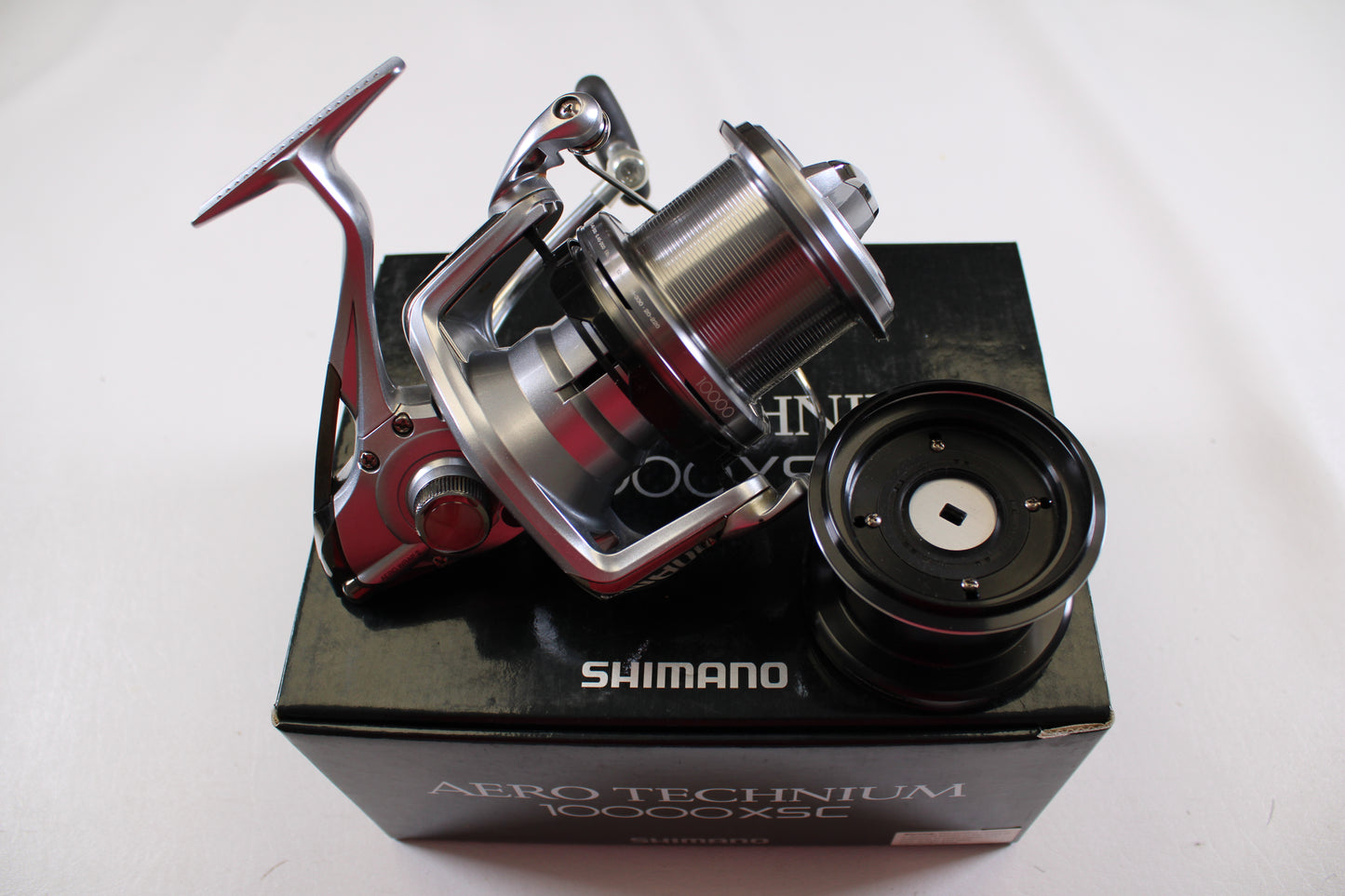 A Shimano Aero Technium 10000XSC Used Spinning Reel in excellent condition rests on its black box with an extra spool beside it, all set against a plain white background.