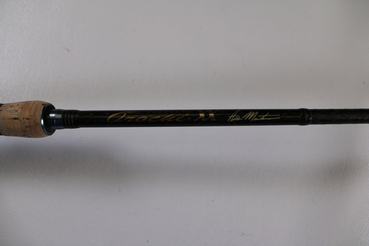 A close-up of the Megabass Orochi XX Aaron Martens F4.1/2-68XX Jerkbait Special used casting rod handle with cork grip and gold signature on a black shaft, ideal for Rod and Reel Trade-In seekers or quality used gear. Plain white background.