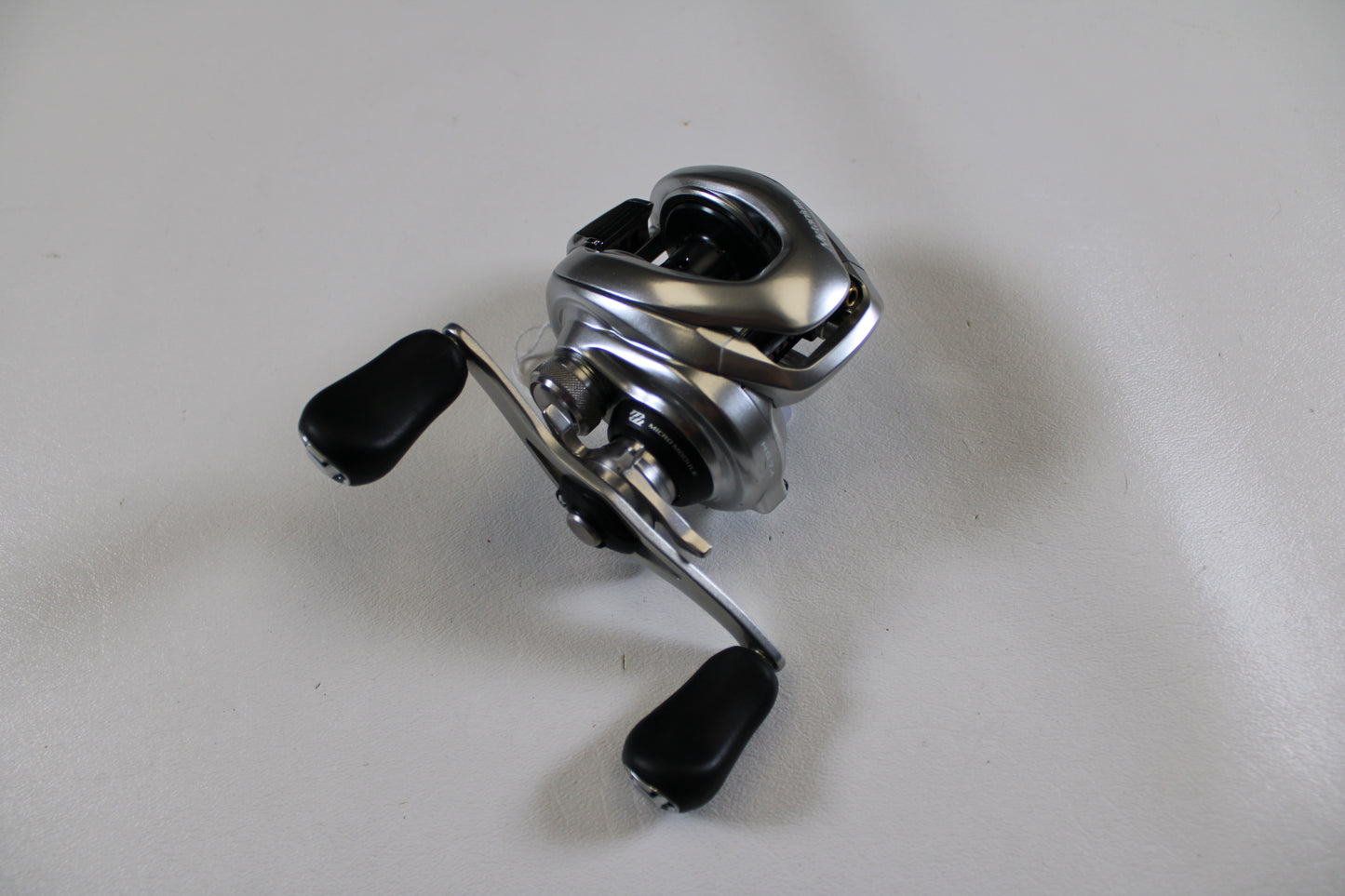A Shimano Metanium MGL 150 6.2:1 Used Casting Reel in very good condition with black handles sits on a white surface—ideal for upgrading via our Rod and Reel Trade-In Program and earning store credit for used gear.