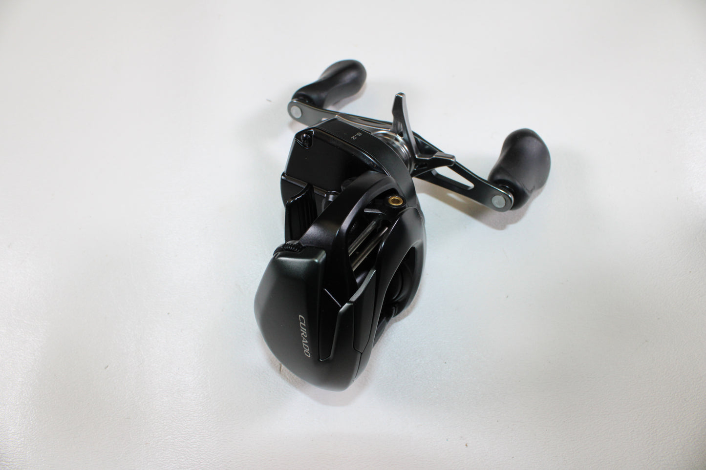 A Shimano Curado CUMGL150 used casting reel in excellent condition, featuring dual handles and a black finish, is displayed on a white surface and viewed from above.