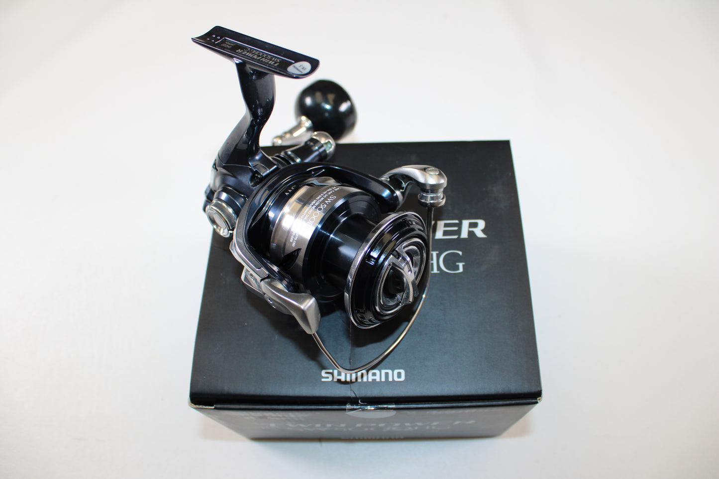 A Shimano Twinpower SW C TPSW5000HGC used spinning reel in mint condition sits atop its original black box packaging with white text, set against a plain white background.