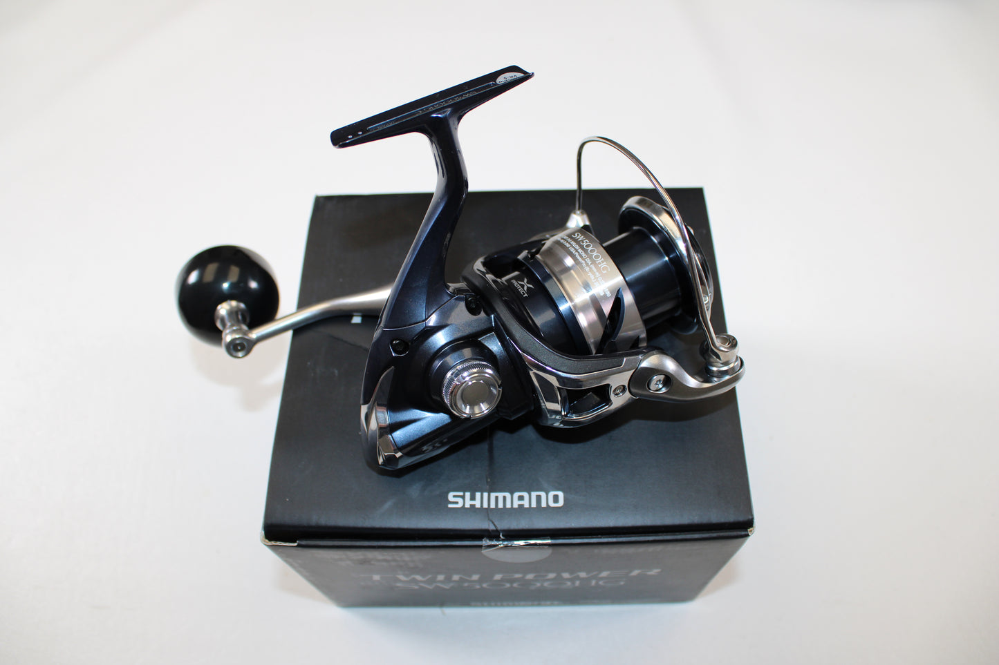 A Shimano Twinpower SW C TPSW5000HGC Used Spinning Reel in mint condition is displayed on its black box with the "Shimano" logo visible, all set against a white background.