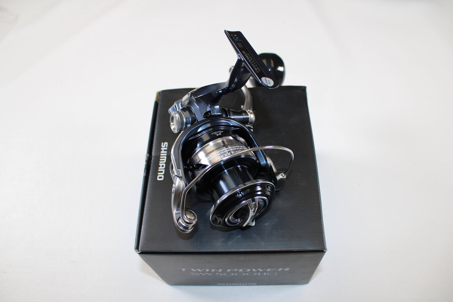 A Shimano Twinpower SW C TPSW8000HGC Used Spinning Reel in mint condition is shown on its black box packaging against a white background, featuring metallic and black details with the Shimano logo visible.