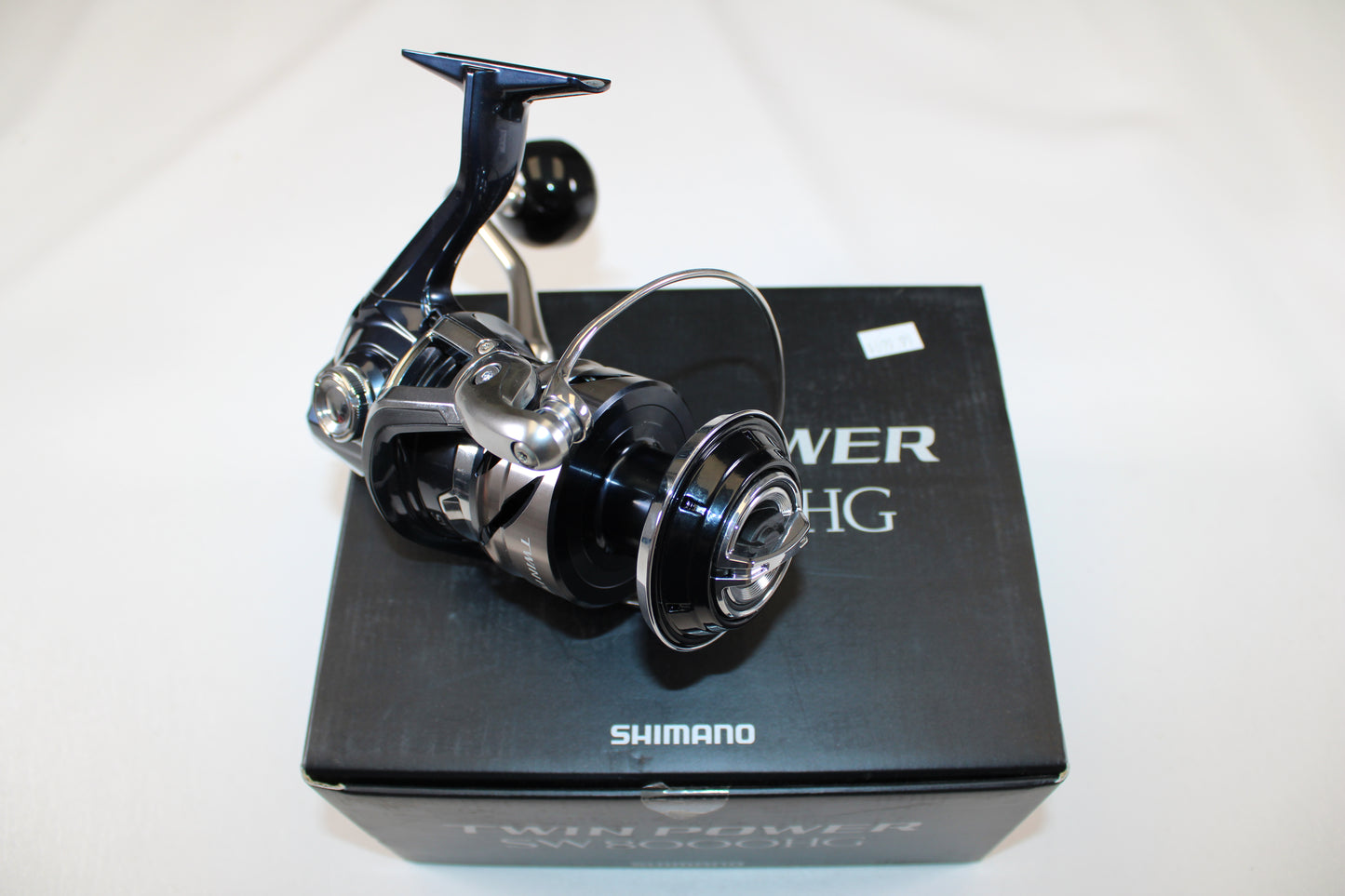A Shimano Twinpower SW C TPSW8000HGC used spinning reel in mint condition sits atop its original black box, featuring visible "Shimano" branding and product details. The reel has a shiny metallic black and silver finish.