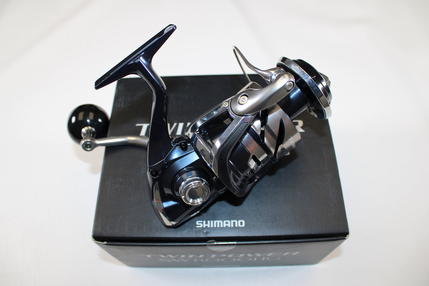 A close-up of a Shimano Twinpower SW C TPSW8000HGC used spinning reel in mint condition, resting on its original black box packaging with the Shimano brand name clearly visible. The reel has a metallic, modern design.