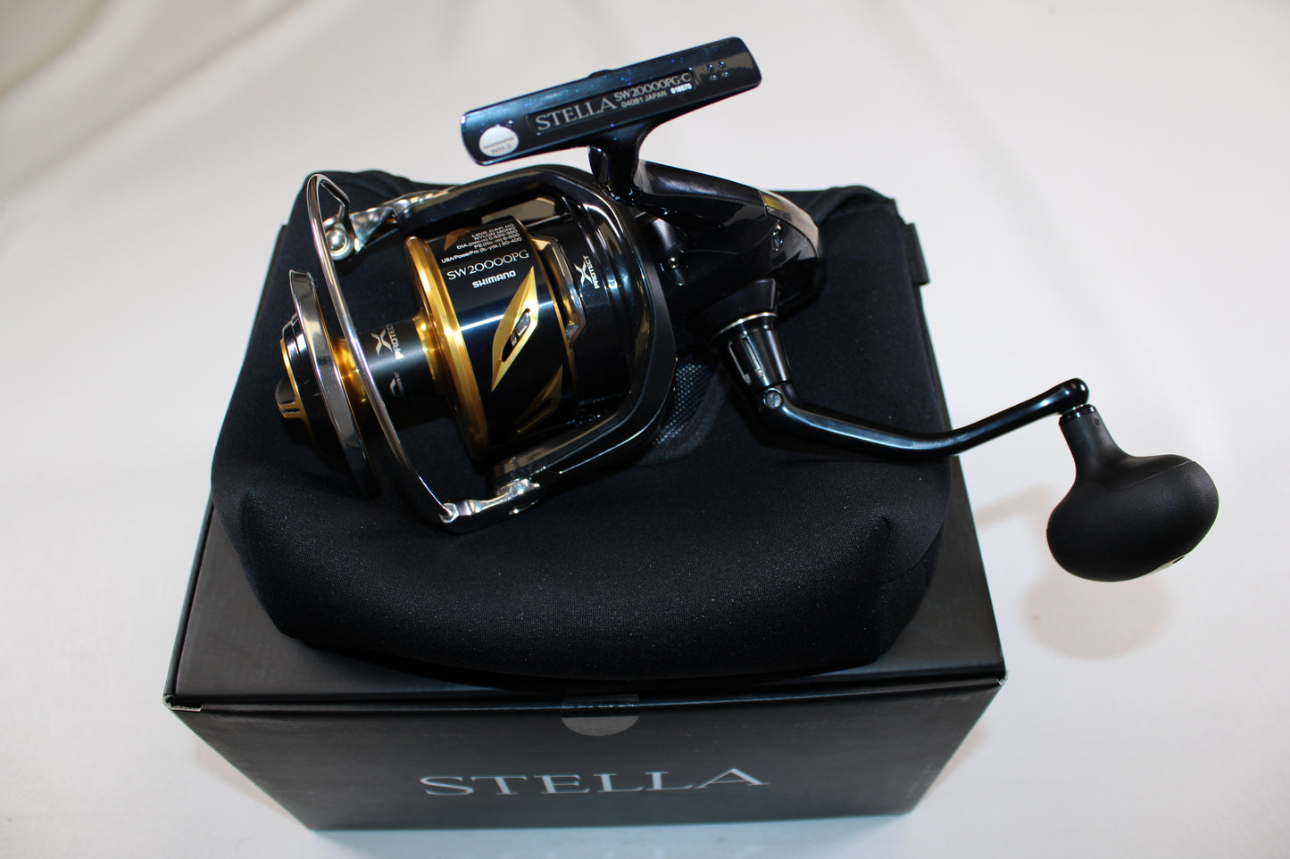 A Shimano Stella SW C STLSW20000PGC spinning reel in mint condition sits on its padded case and "STELLA" box against a white background—ideal for anyone seeking quality used Shimano gear or looking to trade in a rod and reel for store credit.