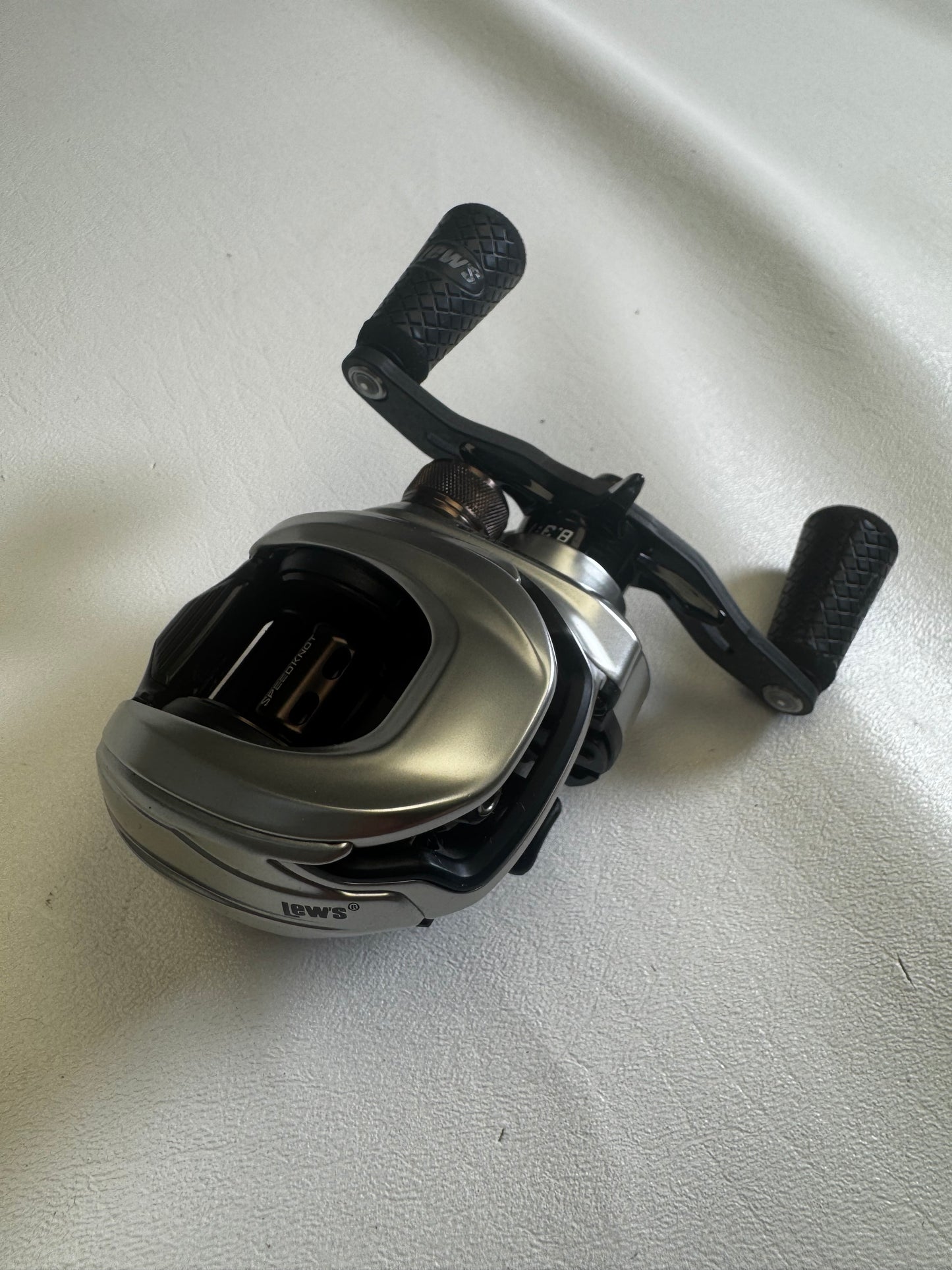 A Lew's HyperMag TLH1XHLG2 Casting Reel, featuring a silver and black design with dual textured handles, sits on a light gray surface. This used reel, in excellent condition, is ideal for upgrading through our Rod and Reel Trade-In program with valuable store credit options.