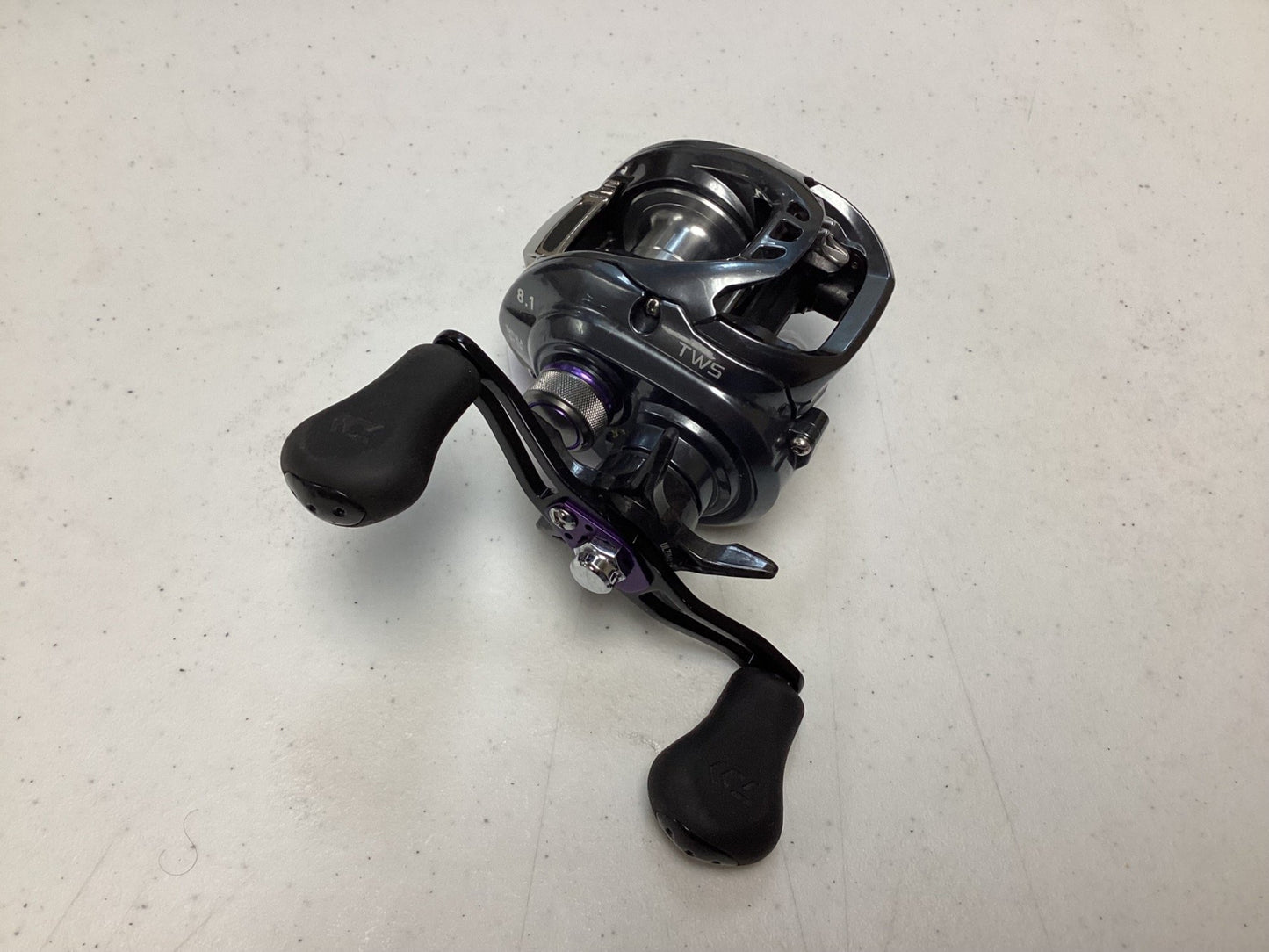 A Daiwa Tatula SV TWS TASV103XS casting reel in black and silver with two large black handles is shown on a white speckled surface. The used reel is in very good condition and made by the brand Daiwa.