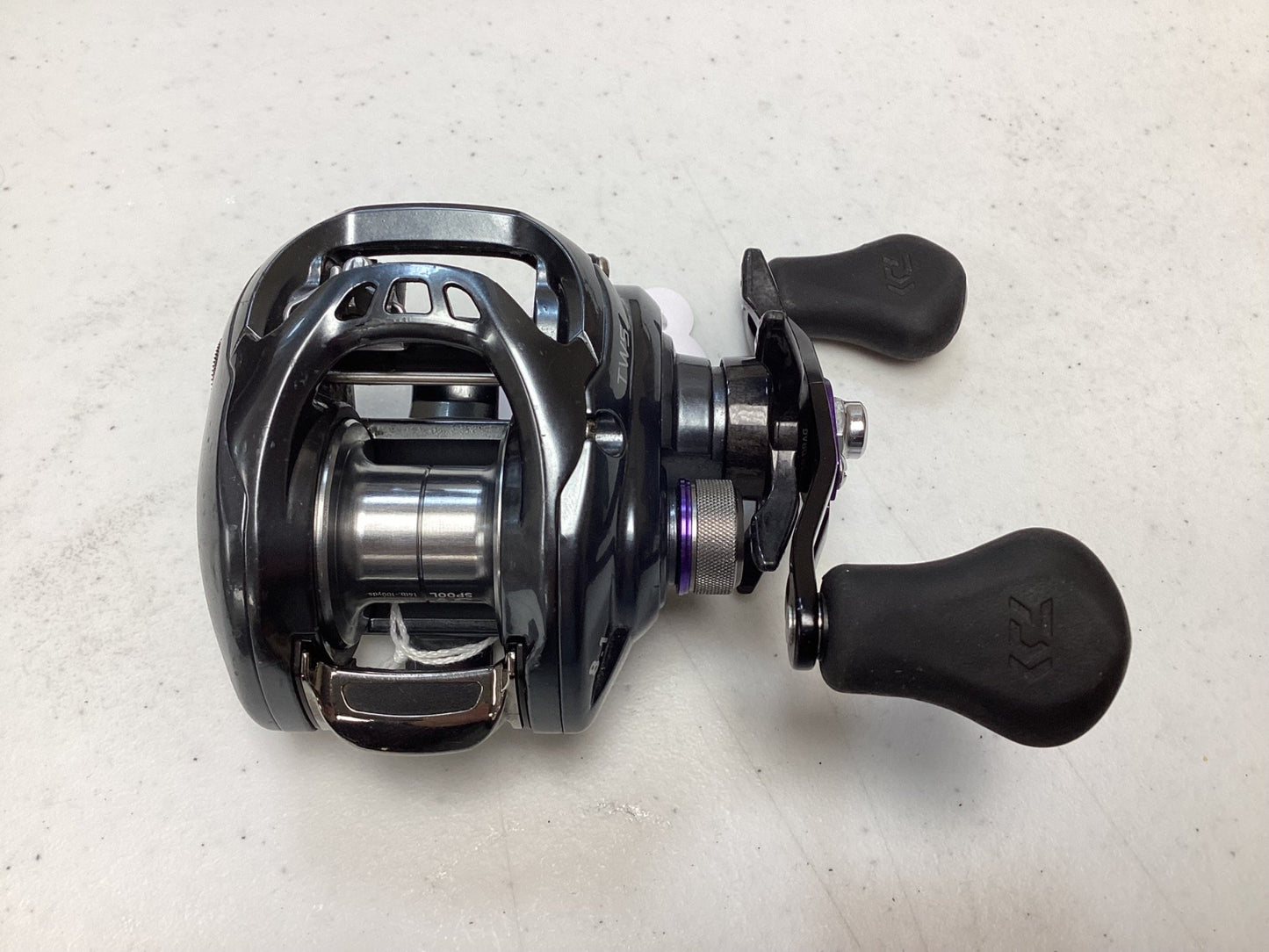 A close-up of a Daiwa Tatula SV TWS TASV103XS used casting reel in very good condition, featuring black and silver design with two black handles, shown on a white textured surface.