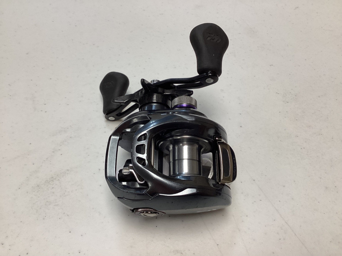 A Daiwa Tatula SV TWS TASV103XS used casting reel in black and silver with dual handles is shown on a white surface; it’s in very good condition.