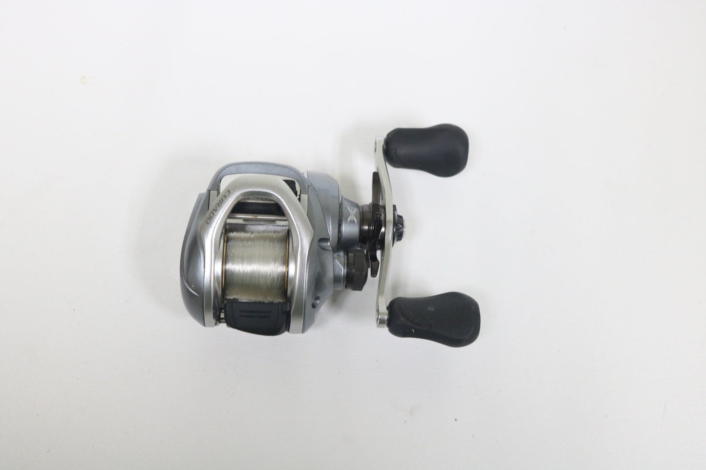 The Shimano Curado I 200PG 5.5:1 Used Casting Reel in excellent condition features a silver design with black handles and spooled line, ideal for anglers upgrading via our Rod and Reel Trade-In or shopping quality Used Gear.