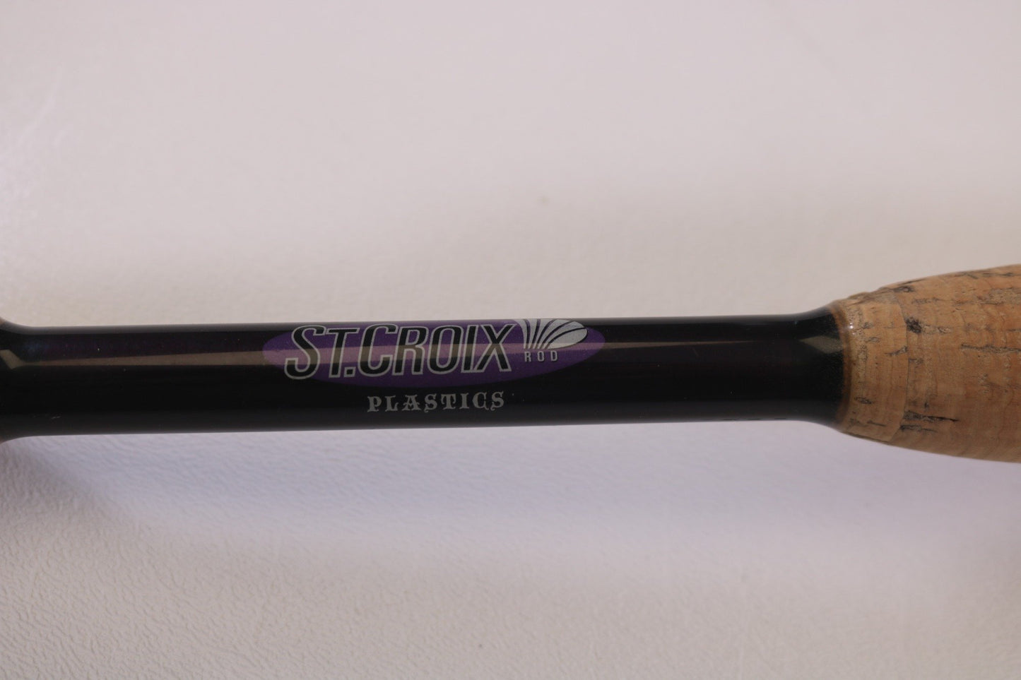 Close-up of a St. Croix Mojo Bass MJC71MF Plastics 7'1" Medium rod handle with "ST. CROIX PLASTICS" visible, part of our Rod and Reel Trade-In Program. The rod boasts a black shaft and cork grip, with text in purple and white on a white background.