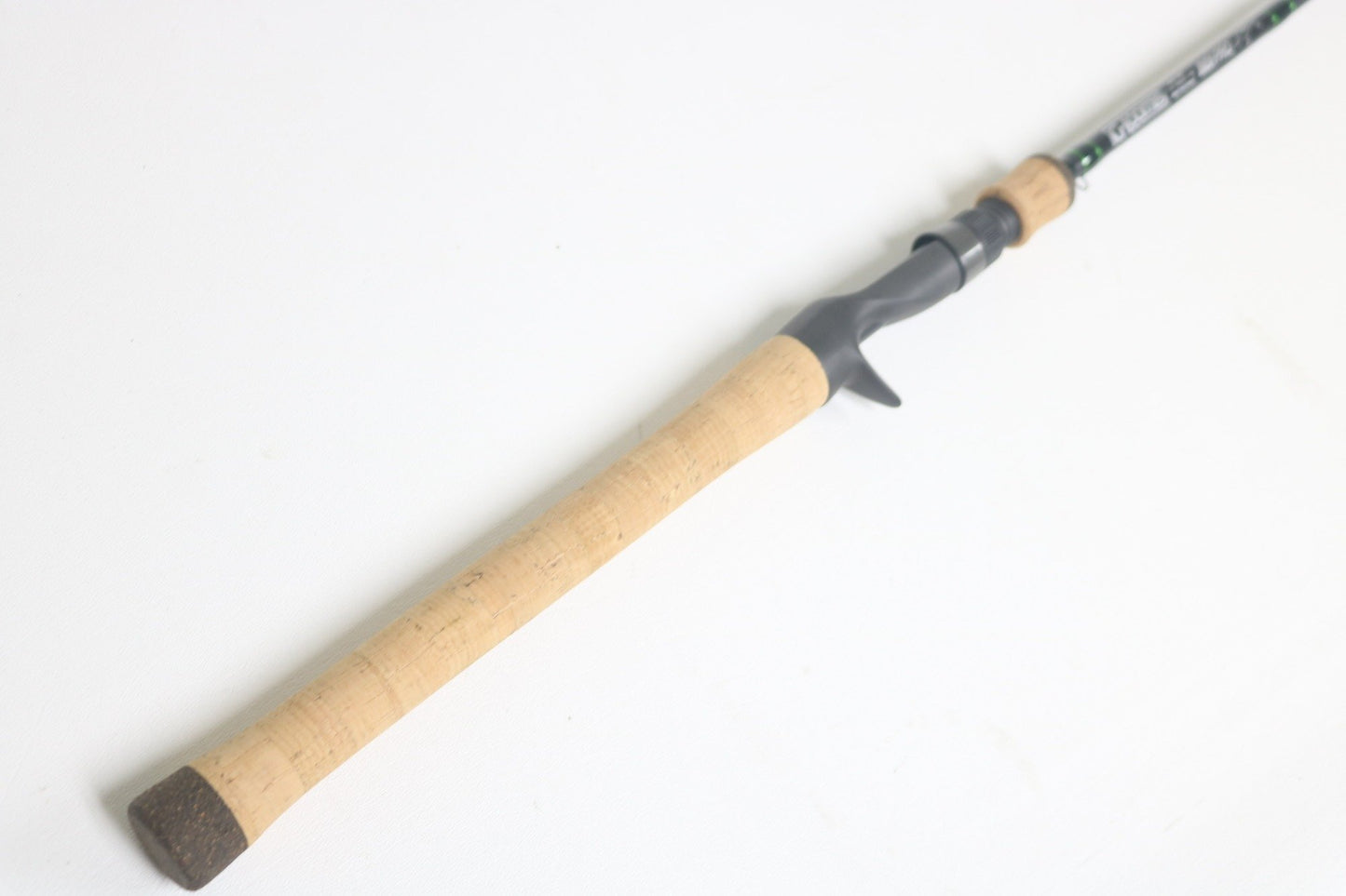 The G. Loomis IMX-Pro 843C CBR Used Casting Rod, partly visible and highlighting the cork handle and grey reel seat, lies diagonally on a white background.