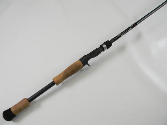 The Kistler KLX KLX-TCJ-70MH 7'0" Medium Heavy used casting rod in excellent condition, part of our Rod and Reel Trade-In Program, rests elegantly on a white surface. It features a sleek black design with a cork handle.