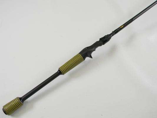 The used Cashion F90474b 7'4" Medium Heavy Casting Rod in very good condition lies on a white surface, featuring a black design with a yellow and black pattern on the handle and butt end, perfect for your next fishing adventure or to trade in for store credit.
