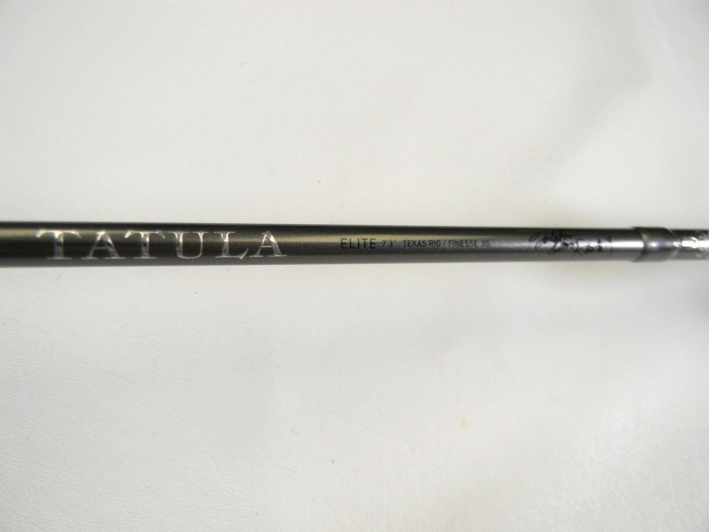 Close-up of a black Daiwa Tatula Elite TTEL731HFB Texas Rig/Finesse Jig 7'3" Heavy rod with "TATULA" and "ELITE" printed in white. The smooth, light backdrop highlights its craftsmanship, making it perfect for our Rod and Reel Trade-In Program for those upgrading used gear with store credit.