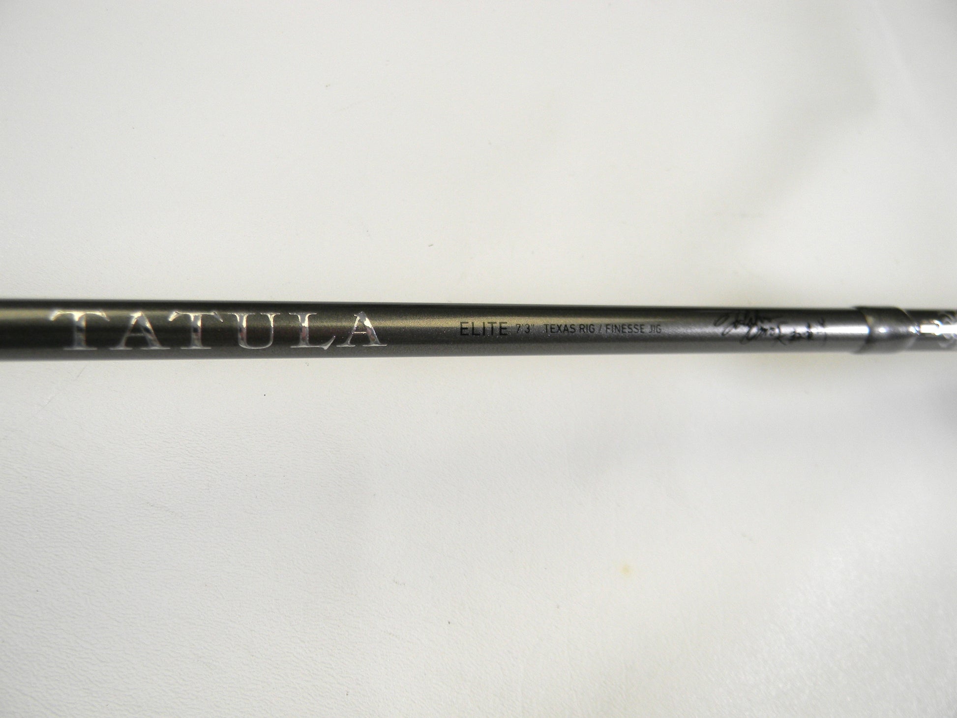 Close-up of a black Daiwa Tatula Elite TTEL731HFB Texas Rig/Finesse Jig 7'3" Heavy rod with "TATULA" and "ELITE" printed in white. The smooth, light backdrop highlights its craftsmanship, making it perfect for our Rod and Reel Trade-In Program for those upgrading used gear with store credit.