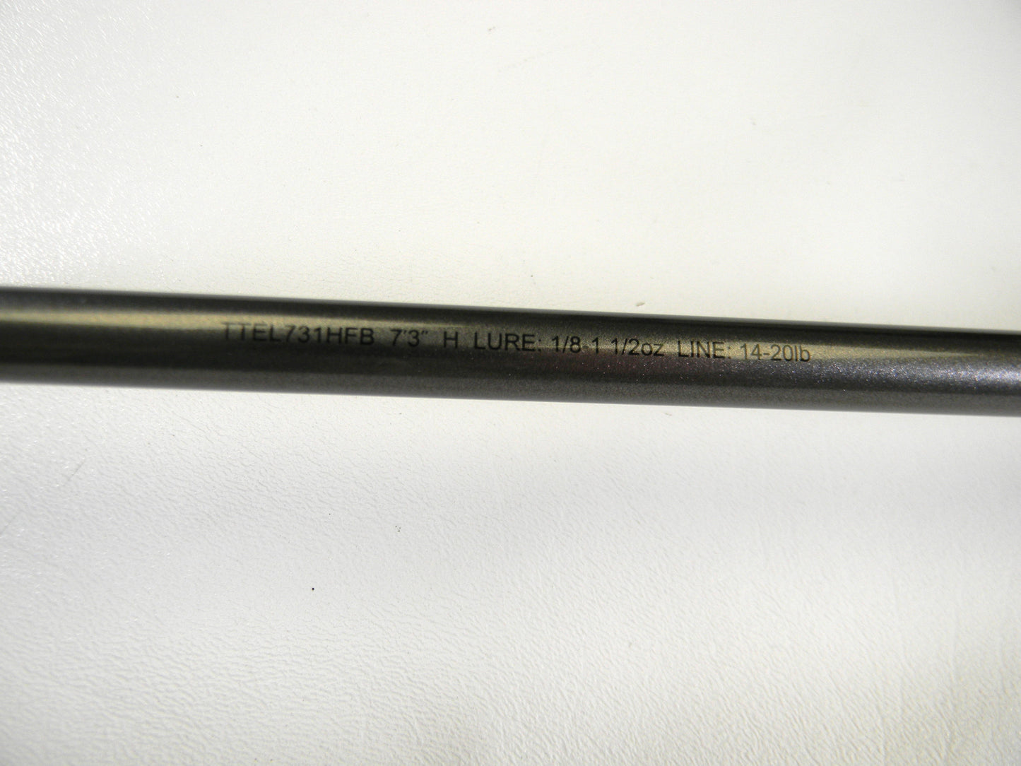 A close-up of the Daiwa Tatula Elite TTEL731HFB Texas Rig/Finesse Jig casting rod, ideal for our Rod and Reel Trade-In Program, shows text on its shaft: "TTEL731HFB 7'3\" H LURE 1/8-1 1/2oz LINE 14-20lb." The mint-condition rod is resting on a white surface.