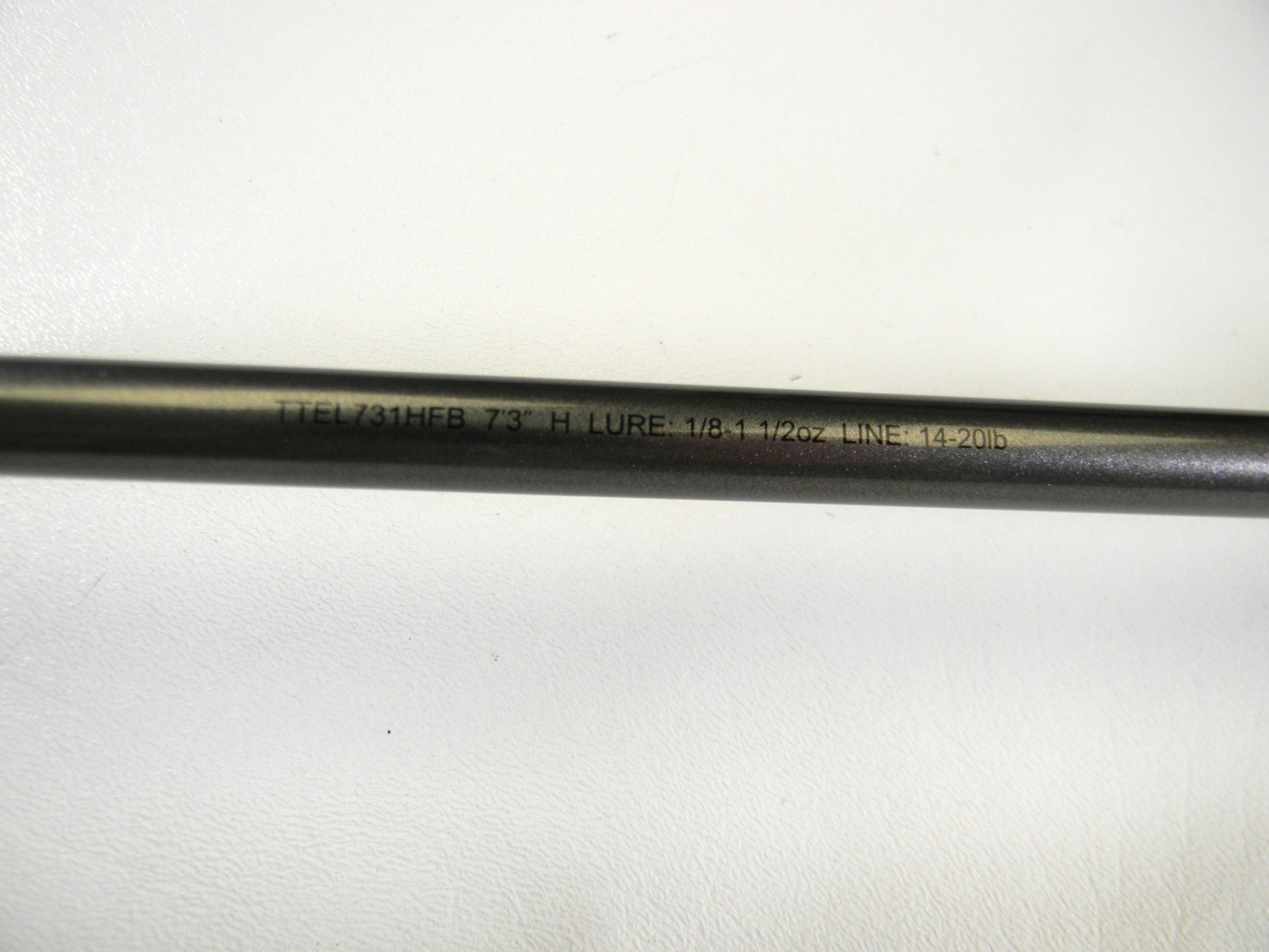 A close-up of the Daiwa Tatula Elite TTEL731HFB Texas Rig/Finesse Jig casting rod, ideal for our Rod and Reel Trade-In Program, shows text on its shaft: "TTEL731HFB 7'3\" H LURE 1/8-1 1/2oz LINE 14-20lb." The mint-condition rod is resting on a white surface.