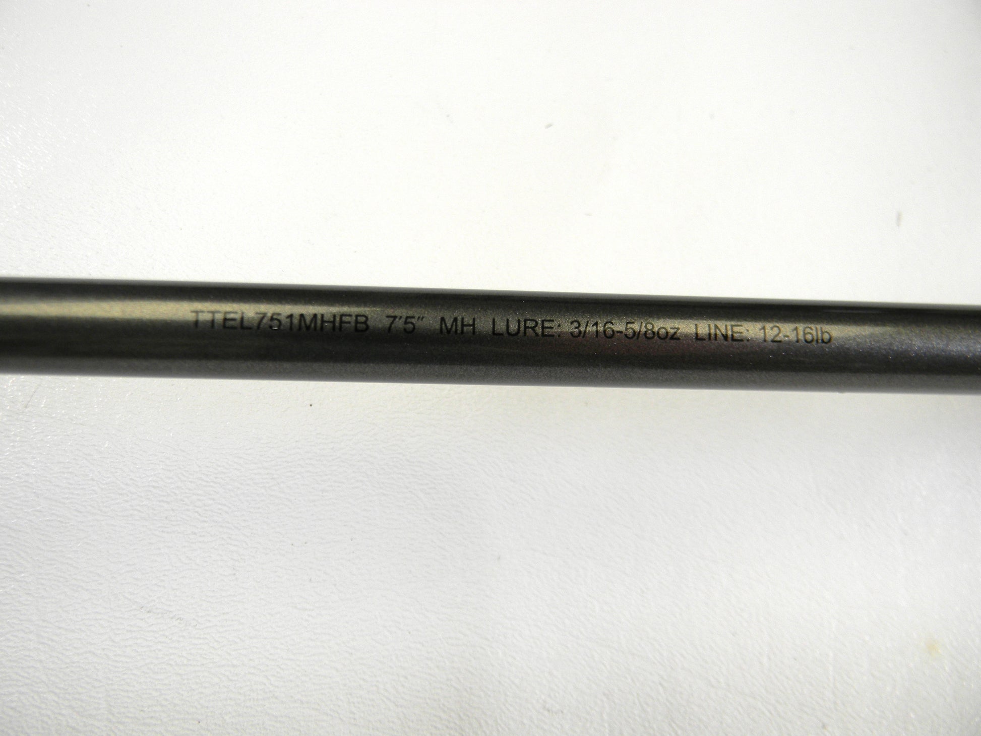 Close-up of a Daiwa Tatula Elite TTEL751MHFB 7'5" MH black fishing rod, with "LURE: 3/16-5/8oz LINE: 12-16lb" engraved. Perfect for upgrading via our Rod and Reel Trade-In Program, converting used gear to store credit. The rod rests on a white surface.