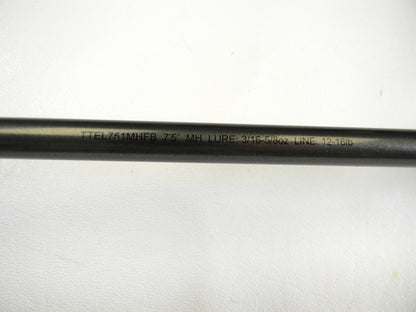 Close-up of a Daiwa Tatula Elite TTEL751MHFB 7'5" MH black fishing rod, with "LURE: 3/16-5/8oz LINE: 12-16lb" engraved. Perfect for upgrading via our Rod and Reel Trade-In Program, converting used gear to store credit. The rod rests on a white surface.