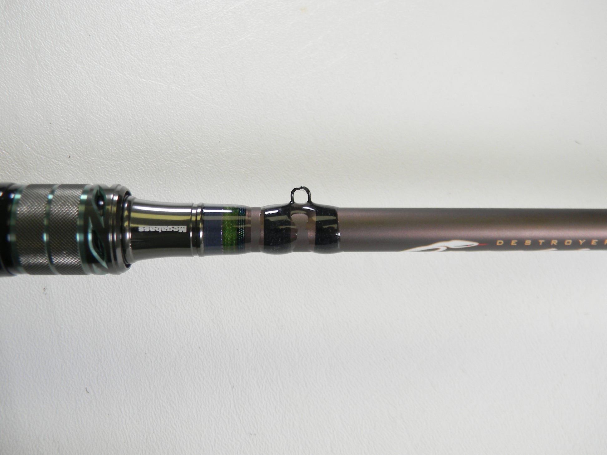 Close-up of a Megabass Orochi X10 F4-68XT Jabberwock 6'8" Medium Light casting rod handle and first line guide against white. The dark, metallic green-accented blank is ideal for our Rod and Reel Trade-In Program if upgrading used gear.