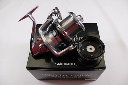 A Shimano Aero Technium 10000XSC Used Spinning Reel in excellent condition rests on its black box with an extra spool beside it, all set against a plain white background.