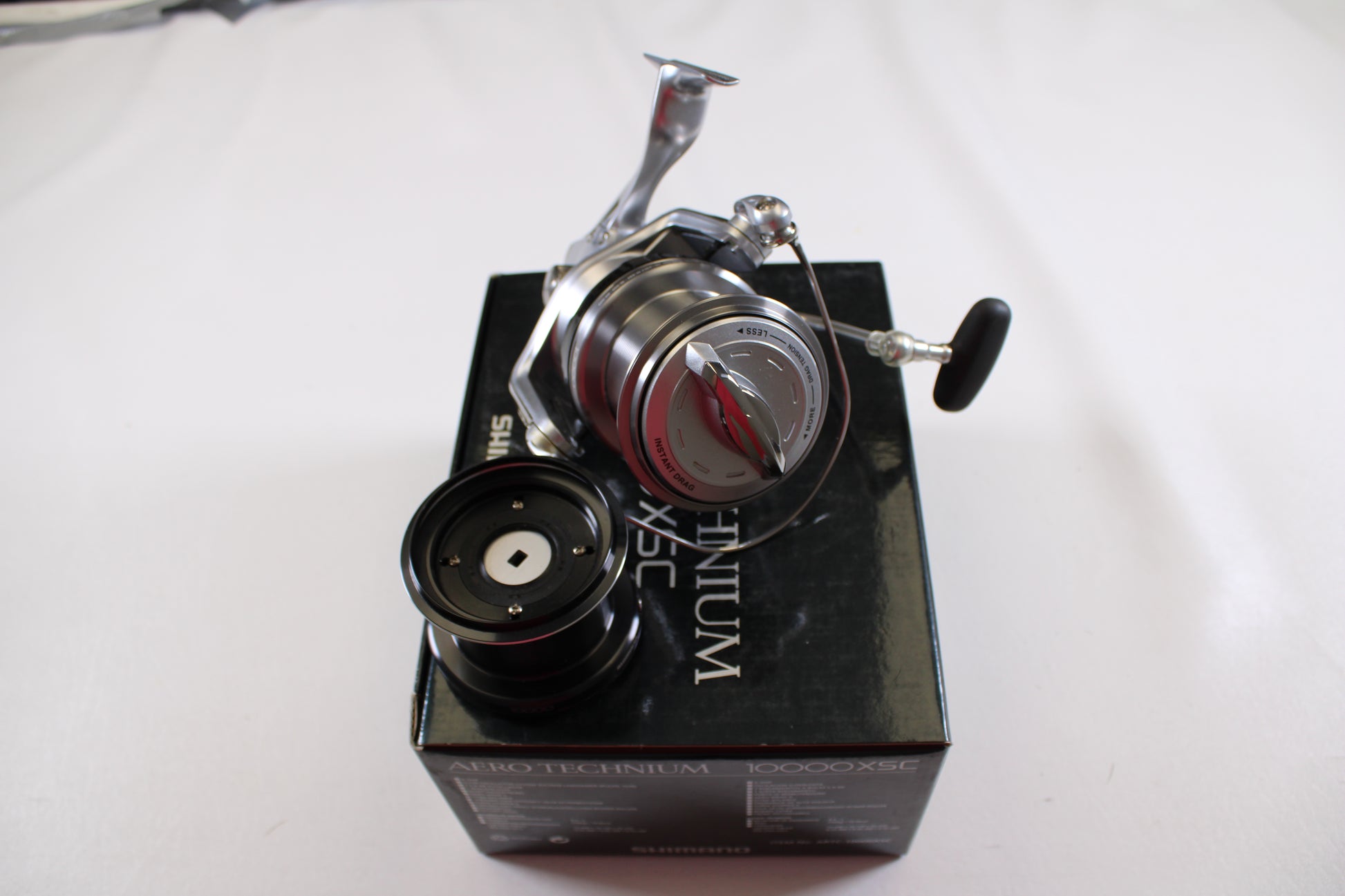 A Shimano Aero Technium 10000XSC used spinning reel in excellent condition with a black handle sits atop a black Shimano box, an extra spool beside it, all arranged on a white surface.
