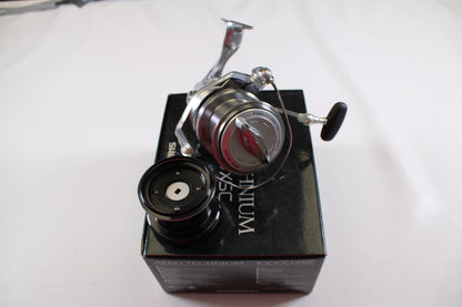 A Shimano Aero Technium 10000XSC used spinning reel in excellent condition with a black handle sits atop a black Shimano box, an extra spool beside it, all arranged on a white surface.