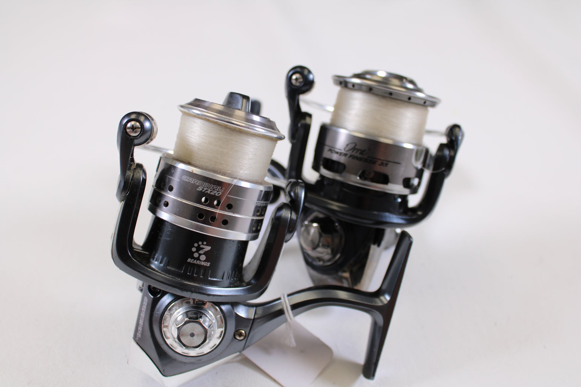 Two Abu Garcia spinning reels—Orra Power Finesse 35 and Cardinal STX20—in black and silver with white line, upright on a white background. Used, good condition; ideal for those interested in deals or our trade-in program. Labels and metallic parts visible.