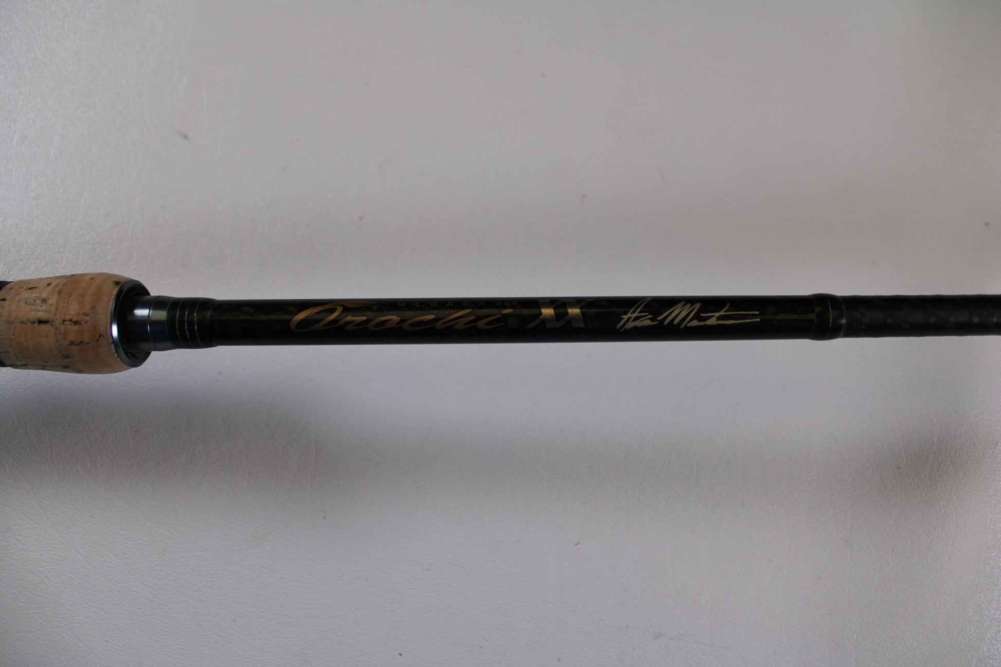 A close-up of the Megabass Orochi XX Aaron Martens F4.1/2-68XX Jerkbait Special used casting rod handle with cork grip and gold signature on a black shaft, ideal for Rod and Reel Trade-In seekers or quality used gear. Plain white background.