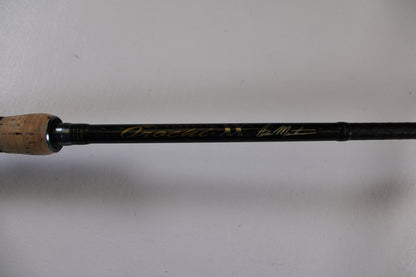 A close-up of the Megabass Orochi XX Aaron Martens F4.1/2-68XX Jerkbait Special used casting rod handle with cork grip and gold signature on a black shaft, ideal for Rod and Reel Trade-In seekers or quality used gear. Plain white background.