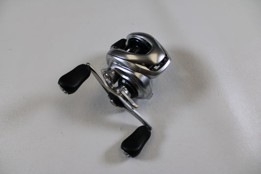 A Shimano Metanium MGL 150 6.2:1 Used Casting Reel in very good condition with black handles sits on a white surface—ideal for upgrading via our Rod and Reel Trade-In Program and earning store credit for used gear.