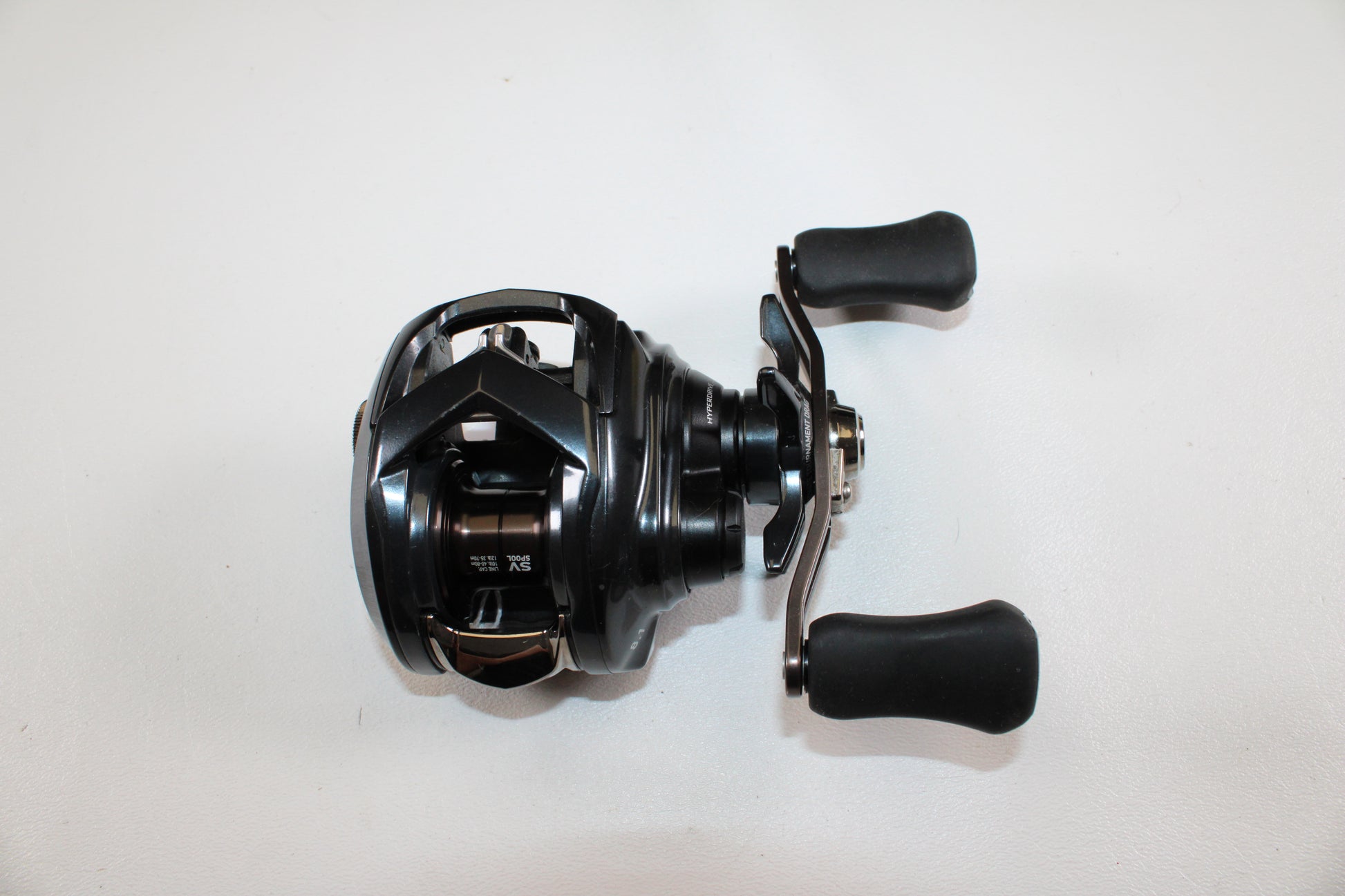 The Daiwa Tatula SV 70 TTUSV70XH Used Casting Reel—black with dual handles and a compact metallic design—is shown on a white background, ideal for trade-ins or store credit through our Rod and Reel Trade-In Program.