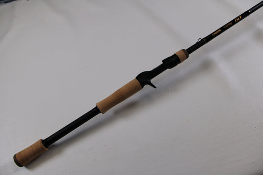 The G. Loomis GLX 895c JWR Used Casting Rod in excellent condition, with cork handles and a trigger grip, lies diagonally on a white surface—ideal for anglers considering our rod and reel trade-in program.