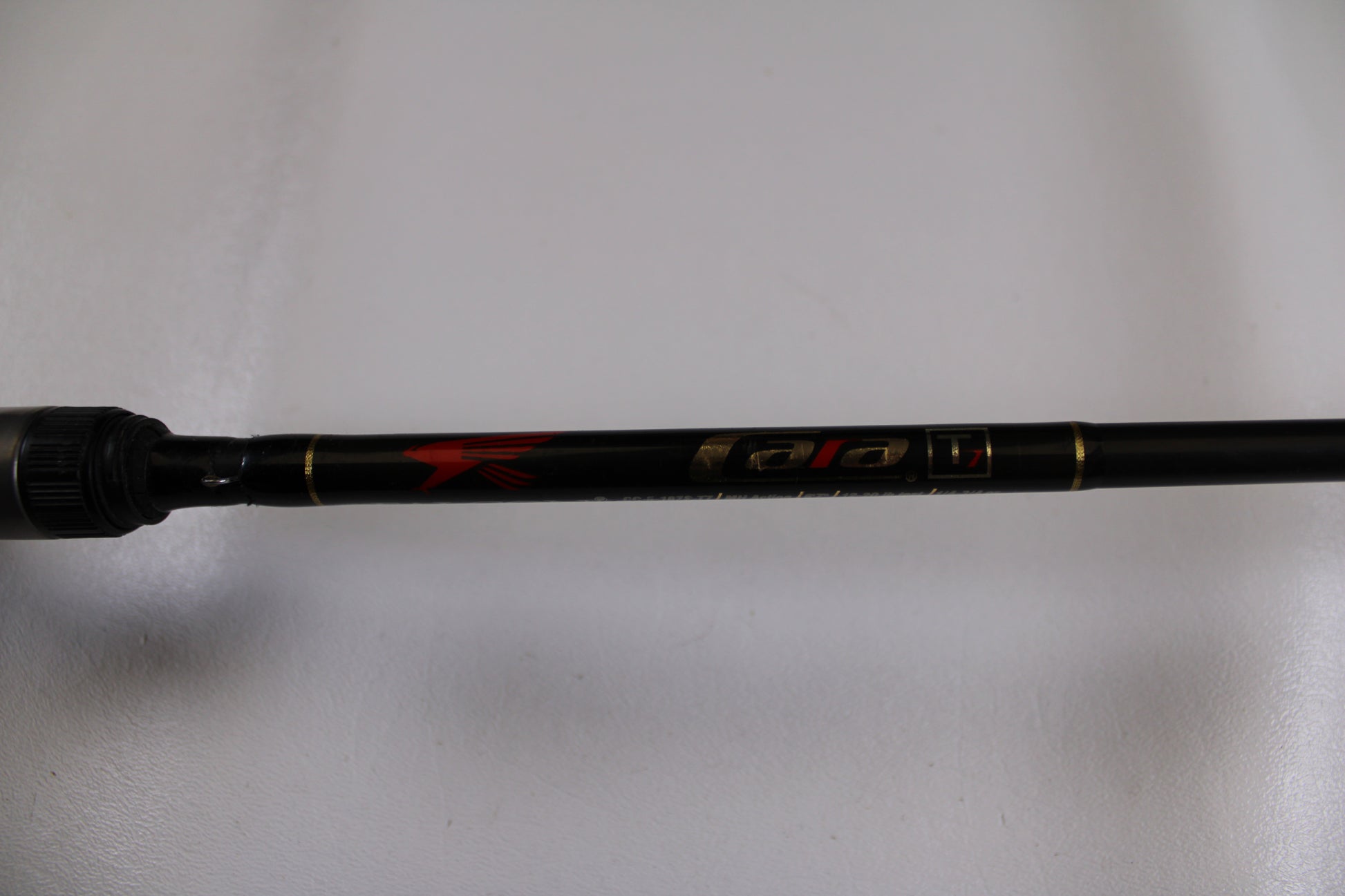 A close-up of a Falcon Cara T7 CC-5-167S Weightless Worm Used Casting Rod in good condition, displaying gold and red accents on black. Trade in your old rod through our program to upgrade and earn store credit.