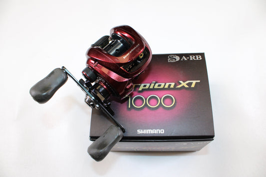 A Shimano Scorpion XT 1000 6.4:1 gear ratio casting reel in very good used condition sits atop its black retail box with gold and white Shimano branding—an excellent choice for quality pre-owned gear or trade-in deals.