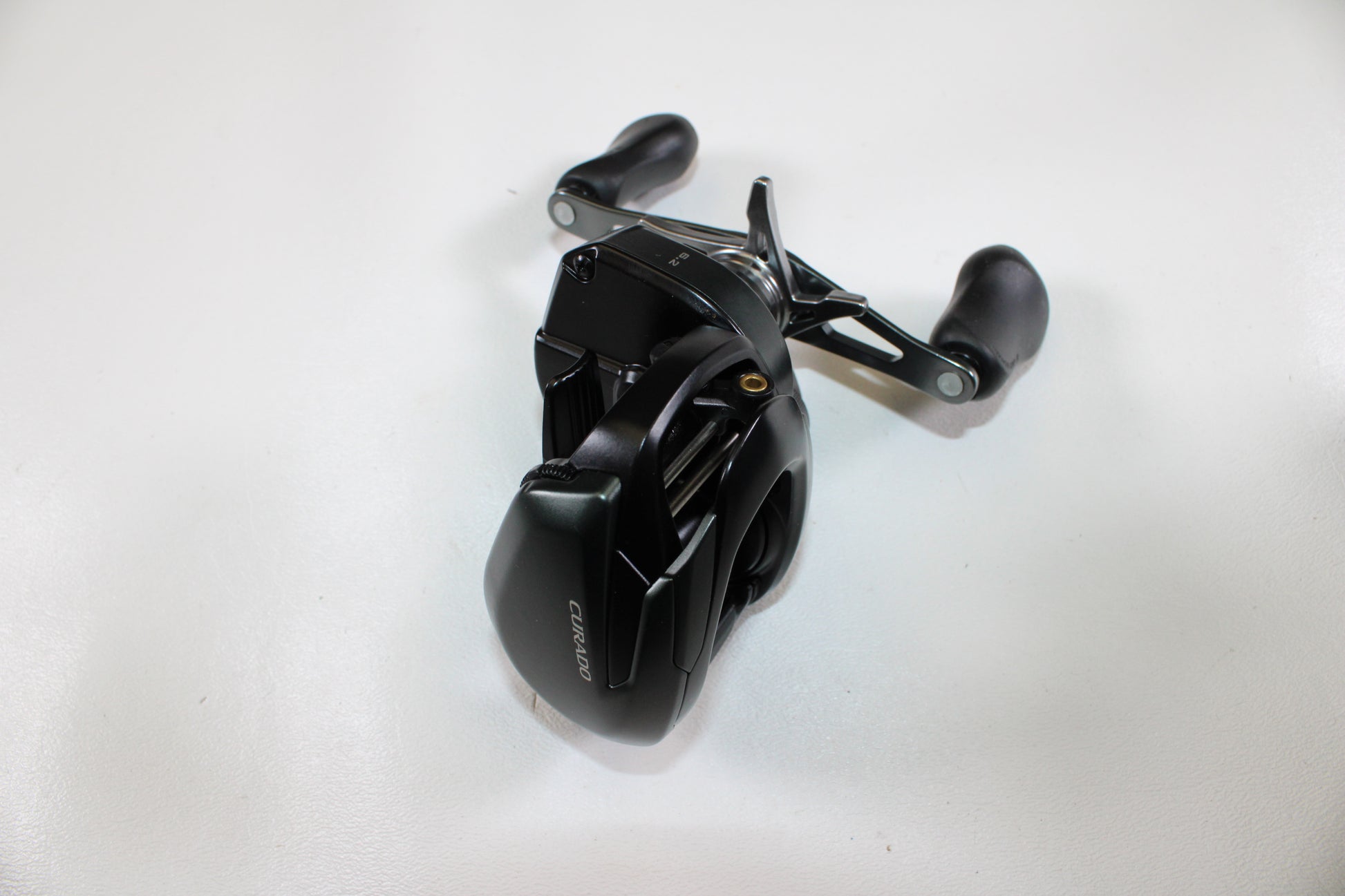 A Shimano Curado CUMGL150 used casting reel in excellent condition, featuring dual handles and a black finish, is displayed on a white surface and viewed from above.