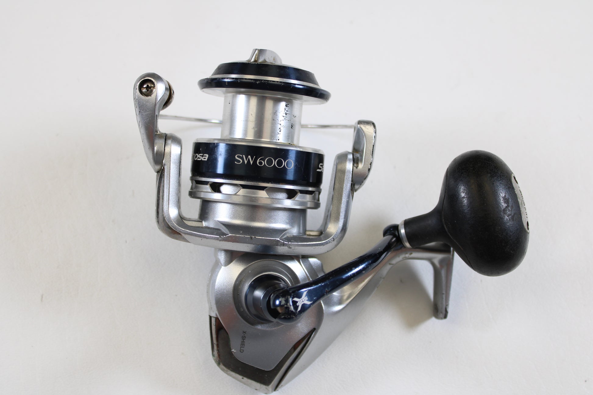 A used Shimano Saragosa SW A SRG6000SWAHG 5.7:1 spinning reel in good condition is shown on a white background. Perfect for anglers upgrading through our Rod and Reel Trade-In Program, it features a large handle and fishing line spool.