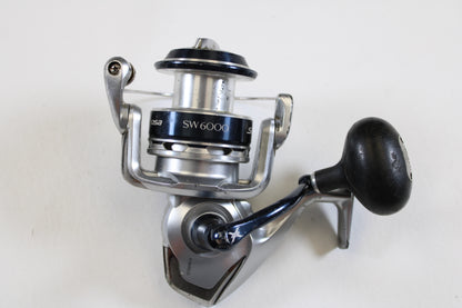 A used Shimano Saragosa SW A SRG6000SWAHG 5.7:1 spinning reel in good condition is shown on a white background. Perfect for anglers upgrading through our Rod and Reel Trade-In Program, it features a large handle and fishing line spool.