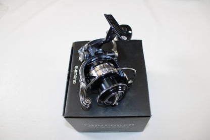 A Shimano Twinpower SW C TPSW5000HGC Used Spinning Reel in mint condition is shown atop its black box packaging against a white background. The reel is metallic with sleek black and silver accents.
