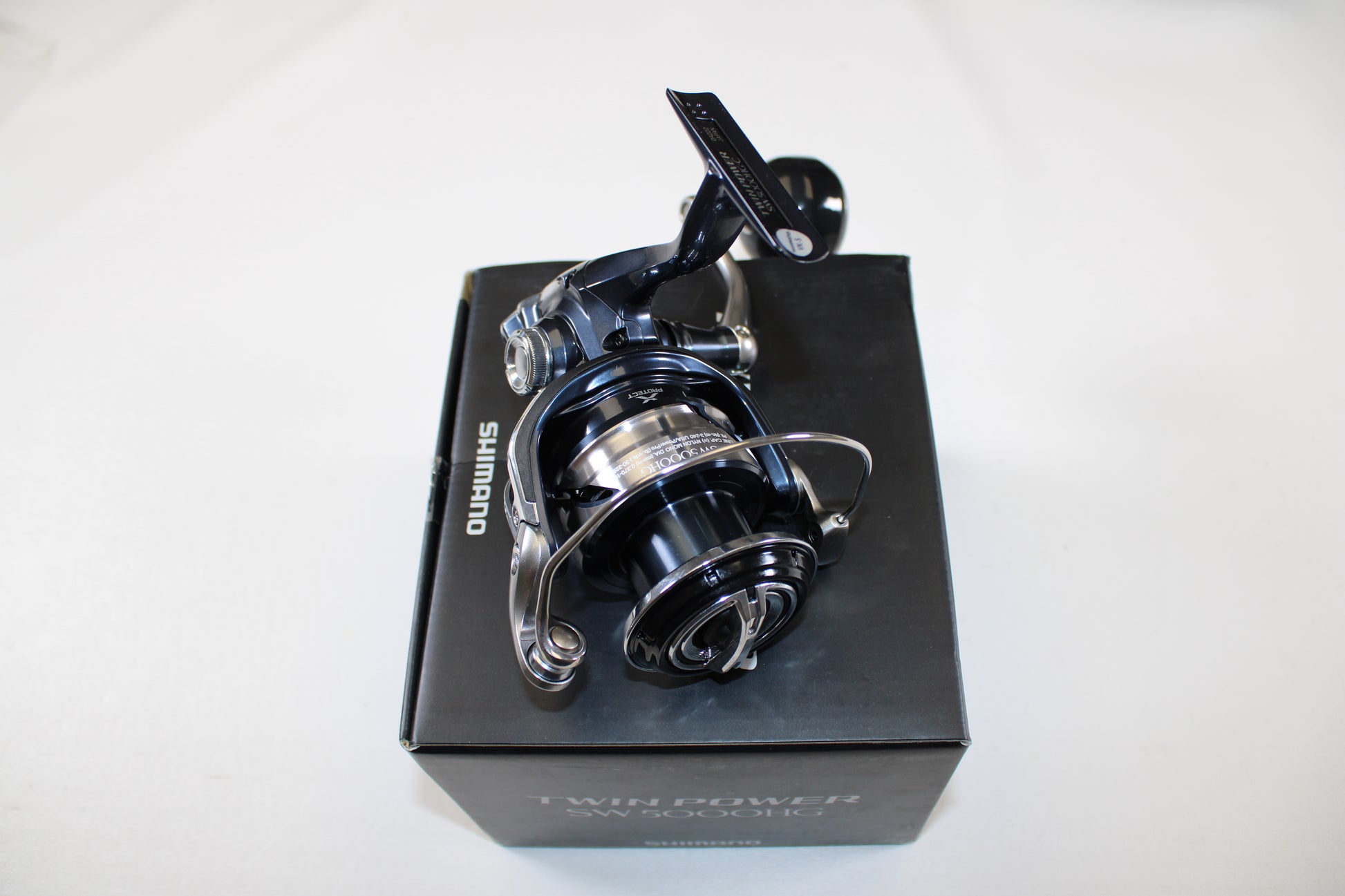 A Shimano Twinpower SW C TPSW8000HGC Used Spinning Reel in mint condition is shown on its black box packaging against a white background, featuring metallic and black details with the Shimano logo visible.