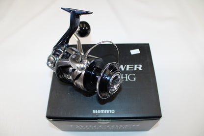 A Shimano Twinpower SW C TPSW8000HGC used spinning reel in mint condition sits atop its original black box, featuring visible "Shimano" branding and product details. The reel has a shiny metallic black and silver finish.