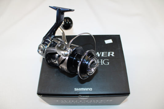 A Shimano Twinpower SW C TPSW8000HGC used spinning reel in mint condition sits atop its original black box, featuring visible "Shimano" branding and product details. The reel has a shiny metallic black and silver finish.