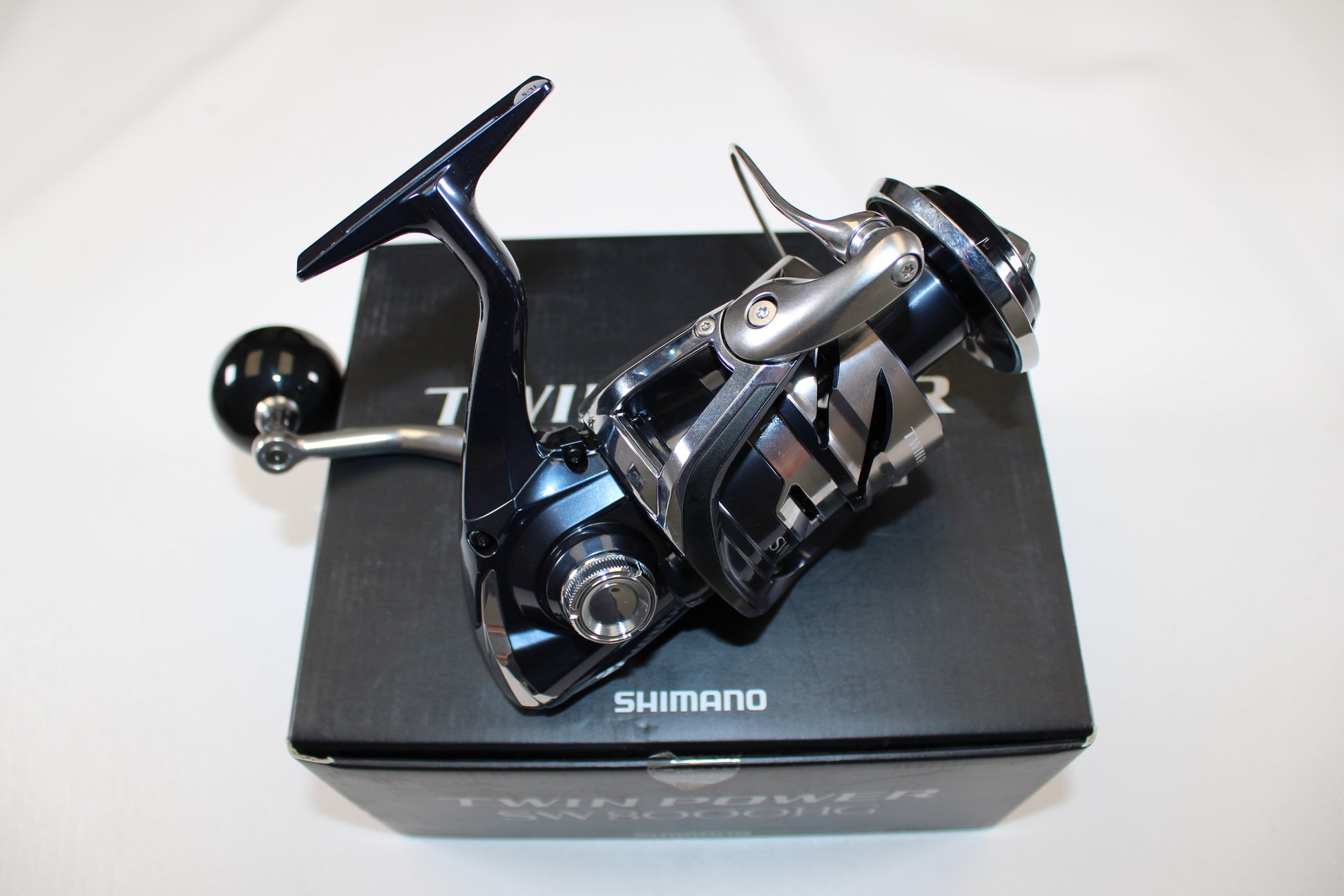 A close-up of a Shimano Twinpower SW C TPSW8000HGC used spinning reel in mint condition, resting on its original black box packaging with the Shimano brand name clearly visible. The reel has a metallic, modern design.