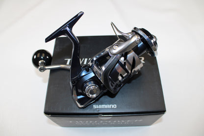 A close-up of a Shimano Twinpower SW C TPSW8000HGC used spinning reel in mint condition, resting on its original black box packaging with the Shimano brand name clearly visible. The reel has a metallic, modern design.