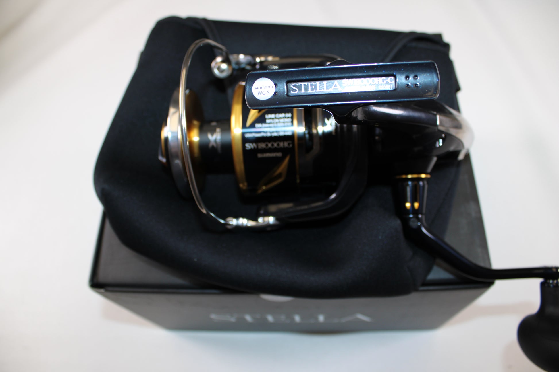 A close-up of a Shimano Stella SW C STLSW18000HGC Used Spinning Reel in mint condition, resting on a soft black pouch atop its box—ideal for those considering our Rod and Reel Trade-In Program for store credit on used Shimano gear.