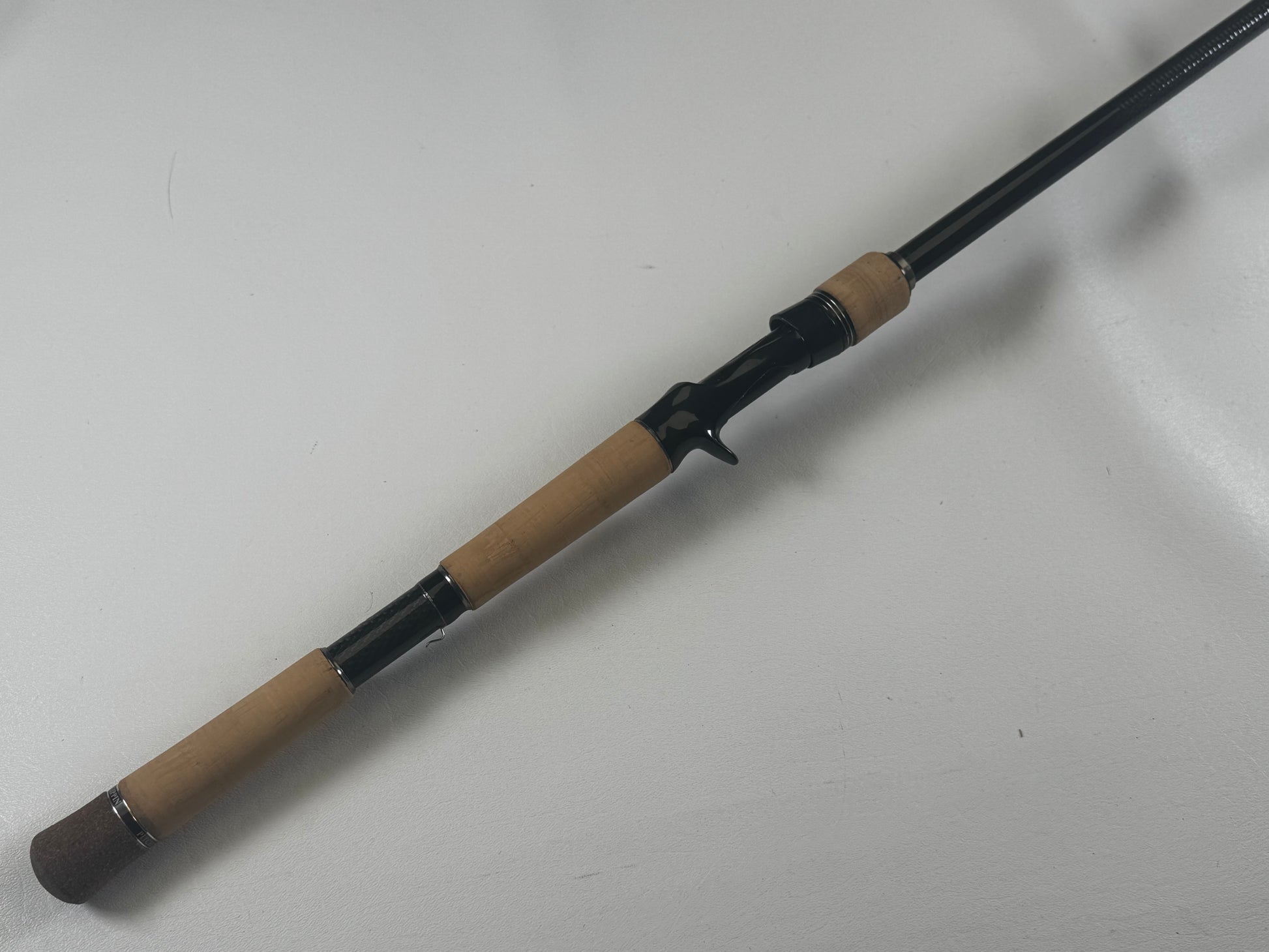 Close-up of a Daiwa BLXSG7111XHXB casting rod on a white surface, showing a cork handle, black reel seat, and the rod extending partially out of frame.