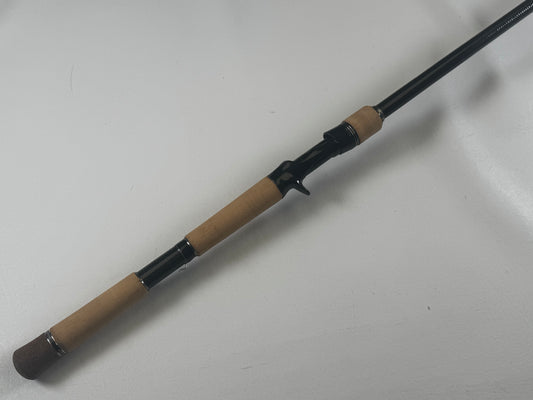 Close-up of a Daiwa BLXSG7111XHXB casting rod on a white surface, showing a cork handle, black reel seat, and the rod extending partially out of frame.